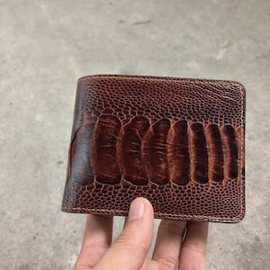 May include: A brown leather wallet with a textured surface and a distinctive pattern. The wallet is held in a hand, highlighting its design and craftsmanship. The wallet appears to be made of high-quality leather.
