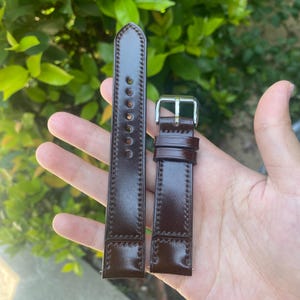 May include: A pair of dark brown leather watch straps with a silver-colored buckle. The straps are held in a hand against a blurred green background. The leather has a glossy finish and visible stitching.