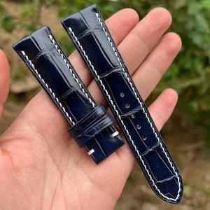 Può includere: Due cinturini blu navy con cuciture bianche. I cinturini hanno una finitura lucida e un motivo strutturato simile alla pelle di coccodrillo. I cinturini sono tenuti in una mano.