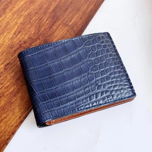 Puede incluir: Una cartera plegable con un exterior azul oscuro y un patrón texturizado de piel de cocodrilo. La cartera tiene bordes visibles de color tostado a lo largo del pliegue inferior, mostrando un contraste entre la superficie azul texturizada y el forro interior liso de color tostado.