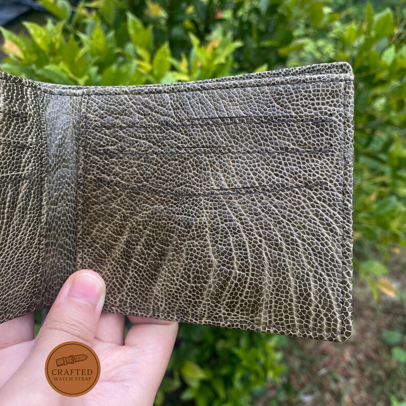 Puede incluir: Cartera de cuero verde oliva abierta con m&uacute;ltiples ranuras para tarjetas. La cartera tiene una superficie texturizada y se mantiene abierta, mostrando el dise&ntilde;o interior. La cartera es de estilo plegable, con un logotipo visible en la parte inferior.