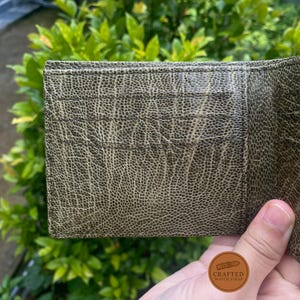 Puede incluir: Primer plano de una cartera de cuero verde oliva texturizado. La cartera tiene m&uacute;ltiples ranuras para tarjetas y una superficie detallada y estampada. El fondo es follaje verde borroso.