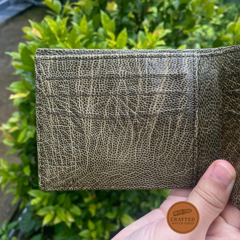 Puede incluir: Primer plano de una cartera de cuero verde oliva texturizado. La cartera tiene m&uacute;ltiples ranuras para tarjetas y una superficie detallada y estampada. El fondo es follaje verde borroso.