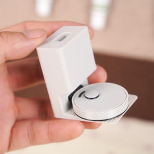 May include: A miniature, light gray robotic vacuum cleaner model. The small, round vacuum has a black accent and is housed in a white charging station. The charging station has a rectangular shape with a slot at the top.