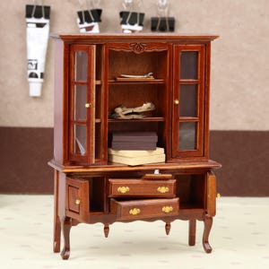 May include: A small, wooden cabinet in a dark brown finish. It features glass-fronted doors and drawers with decorative handles. The cabinet has open doors, revealing shelves and small items inside. The cabinet is a miniature piece.