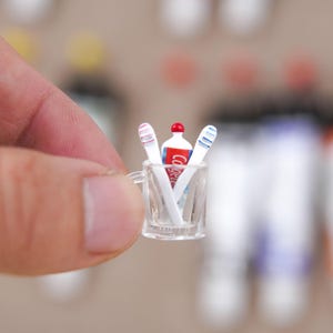 May include: Miniature clear glass cup holding two toothbrushes and a tube of toothpaste. The toothbrushes have red and blue stripes. The toothpaste tube is white with red text that reads "Colgate".