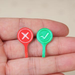 May include: Two small circular tokens with stems, one red with a white X symbol and one green with a white checkmark symbol. These flat, disc-shaped markers feature raised edges and are designed with a distinct color-coded system for indicating.