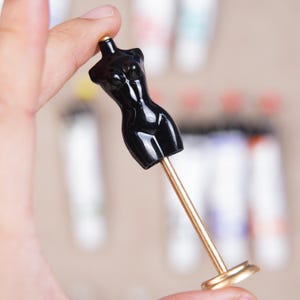 May include: A miniature black dress form with a gold-colored stand and base. The glossy, stylized form is held in a hand, with blurred paint tubes in the background. The dress form is a decorative item.