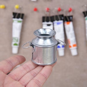 May include: A small, silver metal milk jug with two small handles. The jug is held in a hand, with several tubes of paint in the background. The paint tubes are white with various colored caps.