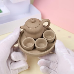 May include: A miniature beige ceramic tea set, including a teapot with a lid, three small cups, and a round serving tray. The set is held by gloved hands, showcasing the delicate craftsmanship and compact size of the tea set.