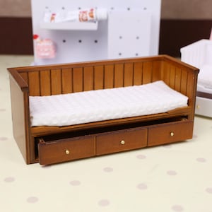 May include: Miniature wooden sofa with three drawers. The sofa has a light brown wooden frame, a white cushion, and three small drawers with gold-coloured knobs. The sofa is designed for a doll's house or miniature setting.
