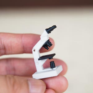 May include: A miniature microscope featuring a white frame and base with black components, including an eyepiece, a revolving nosepiece with multiple objective lenses, a square stage, and a light source housing, all constructed in a compact scale.