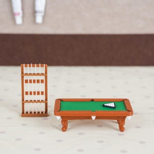 May include: A miniature brown pool table with a green felt surface and a black triangular rack containing white billiard balls, accompanied by a matching brown vertical cue rack holding several thin white pool cues in an upright position.