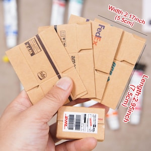 Puede incluir: Una mano sostiene varias cajas de cartón en miniatura, con logotipos de UPS, FedEx y DHL. Las cajas marrones tienen detalles impresos. La caja más grande mide 7,5 cm de largo y 5,5 cm de ancho. El texto incluye 'FRANKFURT'.