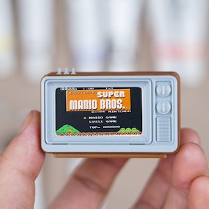 May include: A miniature brown television set featuring a light gray front panel with two circular knobs and a screen displaying the Super Mario Bros. title screen with the text 1986 Nintendo, Mario Game, Luigi Game, and a pixelated landscape.