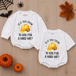 May include: Two white baby bodysuits with long and short sleeves, featuring a graphic of a yellow hard hat and the black text "IS IT TOO SOON TO ASK FOR A HARD HAT?" with small star accents. The garments have snap closures at the bottom.