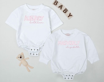 May include: Two white baby onesies with pink text. One says "AUBREY kathleen" and the other says "MAEVERLY elizabeth". A small beige teddy bear toy is next to the onesies. The word "BABY" is spelled out in wooden blocks.