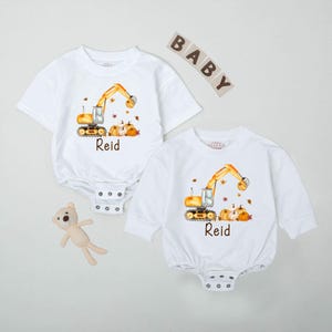 May include: Two white baby bodysuits, one short-sleeved and one long-sleeved, each featuring a yellow excavator design with pumpkins and leaves. The name "Reid" is printed below the design. The word "BABY" is spelled out in wooden letters.