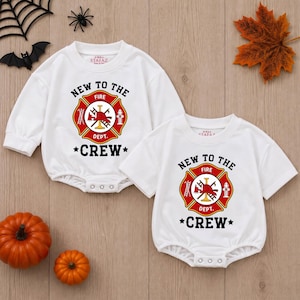 May include: Two white baby bodysuits featuring a red and gold fire department emblem graphic with the text NEW TO THE FIRE DEPT. CREW. One bodysuit has long sleeves and the other has short sleeves, both with three metal snaps at the bottom crotch area.