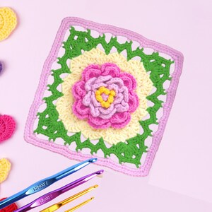 May include: A square, handmade crochet piece featuring a large, pink and yellow flower in the center. The flower is surrounded by green and yellow squares, with a pink border. Crochet hooks in blue, purple, and gold are in the foreground.