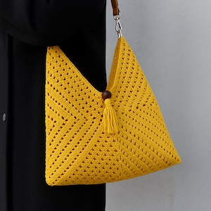 May include: A bright yellow crochet bag with a geometric design. The bag has a brown leather strap and a wooden bead with a yellow tassel. The bag is held by a person wearing a black coat.