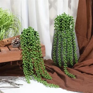 May include: Two artificial string of pearls plants with vibrant green, bead-like foliage cascading downwards. One plant is housed in a textured gray pot, while the other rests on a wooden crate, both featuring long, trailing stems of small spheres.