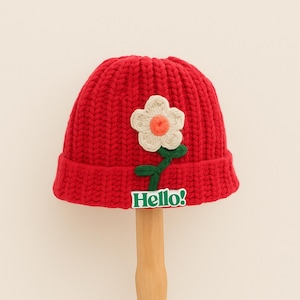 May include: A red knit beanie with a white and orange flower applique and a green stem. The hat has a folded brim and a small green tag that says "Hello!"