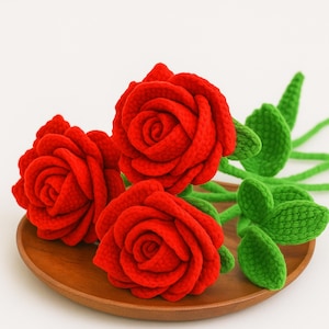 May include: Three handmade red crochet roses with green leaves and stems rest on a wooden plate. The roses have detailed petals and are a vibrant red color. The green leaves and stems provide a natural contrast to the red flowers.