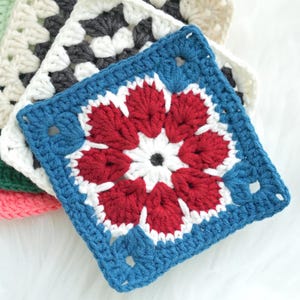 May include: A stack of colorful crochet squares. The top square is blue with a red and white floral design. Other squares include white, black, and green. The squares are made of yarn and are likely handmade.