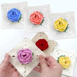 以下が含まれることがあります： 手作りのかぎ針編みの正方形で、カラフルなバラのデザインが特徴です。 バラは青、コーラル、黄色、赤、紫の色合いで、それぞれ緑の葉が付いています。 正方形はオフホワイトで、透かし模様のデザインです。