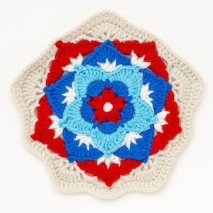 May include: A handmade crochet coaster featuring a floral design. The coaster has a cream-colored border and a central flower pattern in red, light blue, dark blue, and white. The coaster is square with rounded edges.