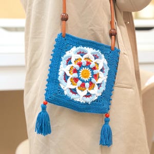 Peut inclure: Sac en crochet bleu carré avec un motif floral central blanc, rouge, jaune et bleu clair. Il est doté d'une bandoulière en cuir marron avec des perles en bois et des pompons bleus.