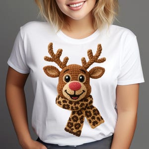 May include: White t-shirt featuring a crocheted reindeer design. The reindeer has brown antlers, a red nose, and a scarf with a brown and tan leopard print. The reindeer's face is detailed with black eyes and a smiling mouth.