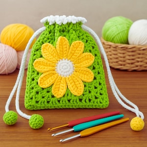 Può includere: Un sacchetto verde all'uncinetto con coulisse e una grande applicazione floreale gialla e bianca. La borsa ha cordini bianchi e pompon verdi. Sono visibili anche tre uncinetti rossi, turchese e gialli.
