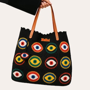 Puede incluir: Bolso tote negro con un diseño de ojos repetido en varios colores. El bolso tiene asas de cuero marrón y la palabra "Hello!" en rojo. El bolso está hecho de un material tejido.