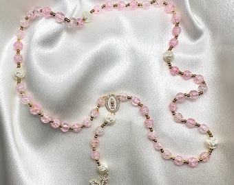 May include: A pink beaded rosary with gold-colored accents and a decorative cross pendant. The rosary features pink, crackled beads, white rose-shaped beads, and a gold-colored cross with clear crystal accents.