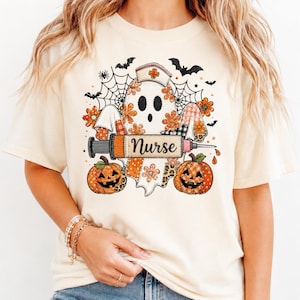 Può includere: Una t-shirt color crema con la grafica di un fantasma che indossa una cuffia da infermiere, circondato da ragnatele, pipistrelli e zucche. Il fantasma tiene una siringa con la scritta Nurse. Il design include motivi floreali e a quadri.