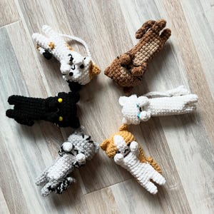 Peut inclure: Six figurines de chats au crochet disposées en cercle. Les chats présentent divers motifs de couleurs, notamment noir uni avec des yeux jaunes, blanc uni avec des yeux bleus, tabby brun, tabby gris, orange et blanc, et blanc avec des taches orange et noires.