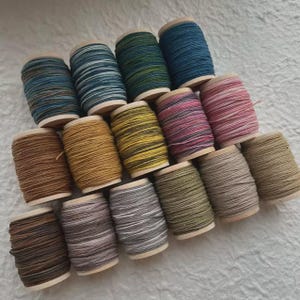 May include: A collection of fifteen wooden spools wrapped in thread of various colors, including shades of blue, green, yellow, pink, brown, and gray. The spools are arranged in a triangular formation, showcasing a diverse palette of textile fibers.