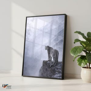 May include: Framed art print depicting a snow leopard on a cliff edge, with a misty mountain backdrop. The black frame encloses the image, which is in monochrome tones. The print is propped against a white wall.