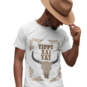 Puede incluir: Camiseta blanca con un gráfico de calavera de toro y el texto "Yippy Kai Yay" en marrón. El diseño está enmarcado por adornos dorados. La persona de la imagen lleva un sombrero de vaquero marrón.