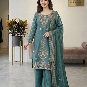 Puede incluir: Conjunto salwar kameez bordado de color turquesa. El atuendo incluye una túnica de manga larga, pantalones de pernera ancha y una bufanda a juego. El bordado es en hilo dorado, y el atuendo lo lleva una persona. El letrero de fondo dice "Featuring Hania Aamir."