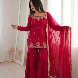 Puede incluir: Un vibrante conjunto de salwar kameez rojo. El atuendo incluye una túnica de manga larga con bordados dorados, pantalones de pierna ancha a juego y una dupatta transparente con ribete dorado. El atuendo lo lleva una persona.