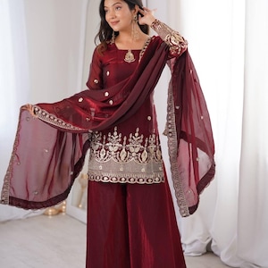 May include: A burgundy three-piece outfit, comprising a long-sleeved tunic with gold embroidery and wide-leg trousers. The ensemble includes a sheer scarf with matching embellishments. The sleeves and scarf edges feature detailed gold embroidery.