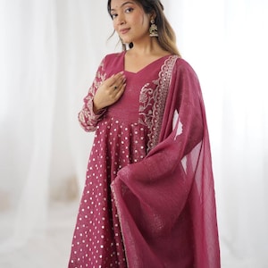 May include: A maroon-colored Indian salwar kameez set. The top features a V-neck, long sleeves with gold embroidery, and a flared silhouette with white polka dots. A matching dupatta and pants complete the ensemble. The outfit is suitable for festive occasions.