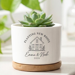 May include: A white ceramic cylindrical planter with a bamboo saucer base containing a green succulent. The planter features a black graphic of a house with the text "PLANTING NEW ROOTS", "Emma & Noah", and the year "2026" printed on the front.