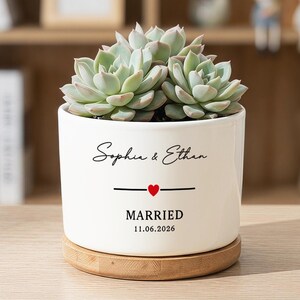 May include: A white ceramic cylindrical planter containing a green succulent plant. The planter features the black text "Sophie & Ethan" above a small red heart, the word "MARRIED", and the date "11.06.2026". It sits on a round wooden base.
