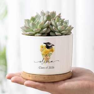 May include: A white ceramic cylindrical planter with a bamboo saucer containing a green succulent. The front features a graphic of yellow flowers topped with a black graduation cap, the name Olivia in script, and the text Class of 2026.