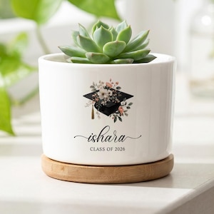 May include: A white ceramic planter with a bamboo base containing a green succulent. The planter features a graphic of a black graduation cap adorned with floral accents, the name "ishara" in script, and the text "CLASS OF 2026" printed below.