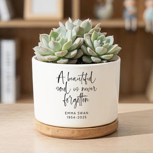 May include: A white ceramic planter with a light wood base containing a green succulent. The planter features black text that reads A beautiful soul is never forgotten, followed by the name Emma Swan and the years 1954-2025 in a clean font.